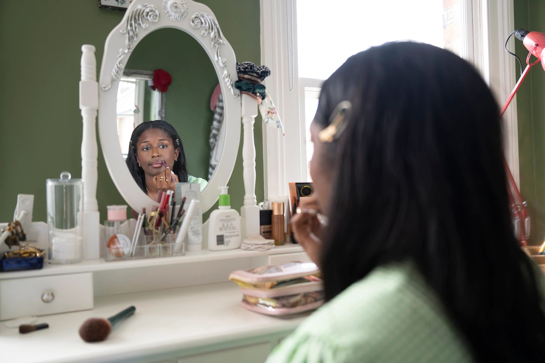 Over the shoulder view of a young woman applying make up in a mirror at home