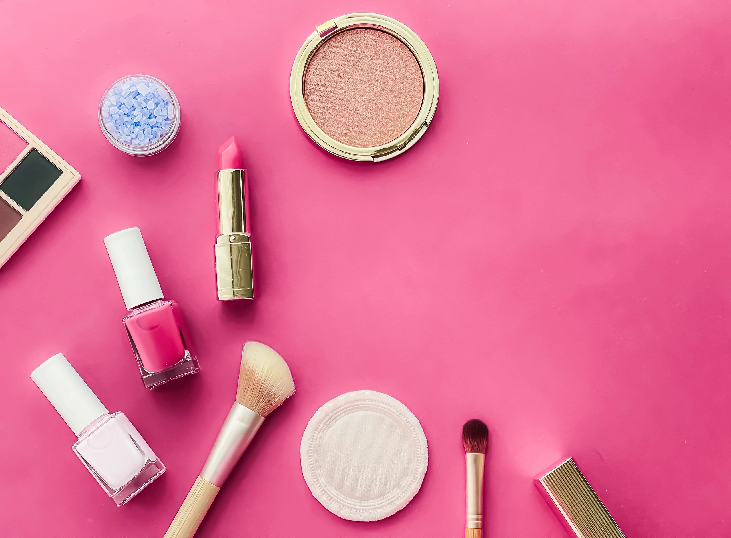 Cosmetic products arranged on a pink background, featuring blush, lipstick, nail polish, and brushes for makeup and beauty care