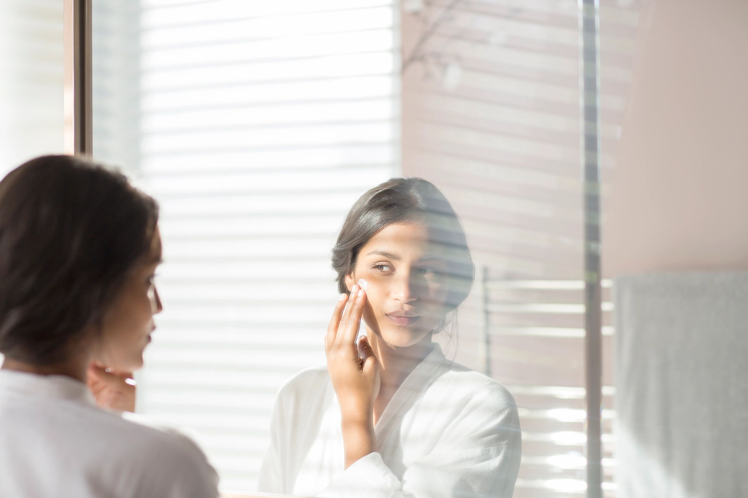 Woman applying moisturizer to cheek in bathroom mirror